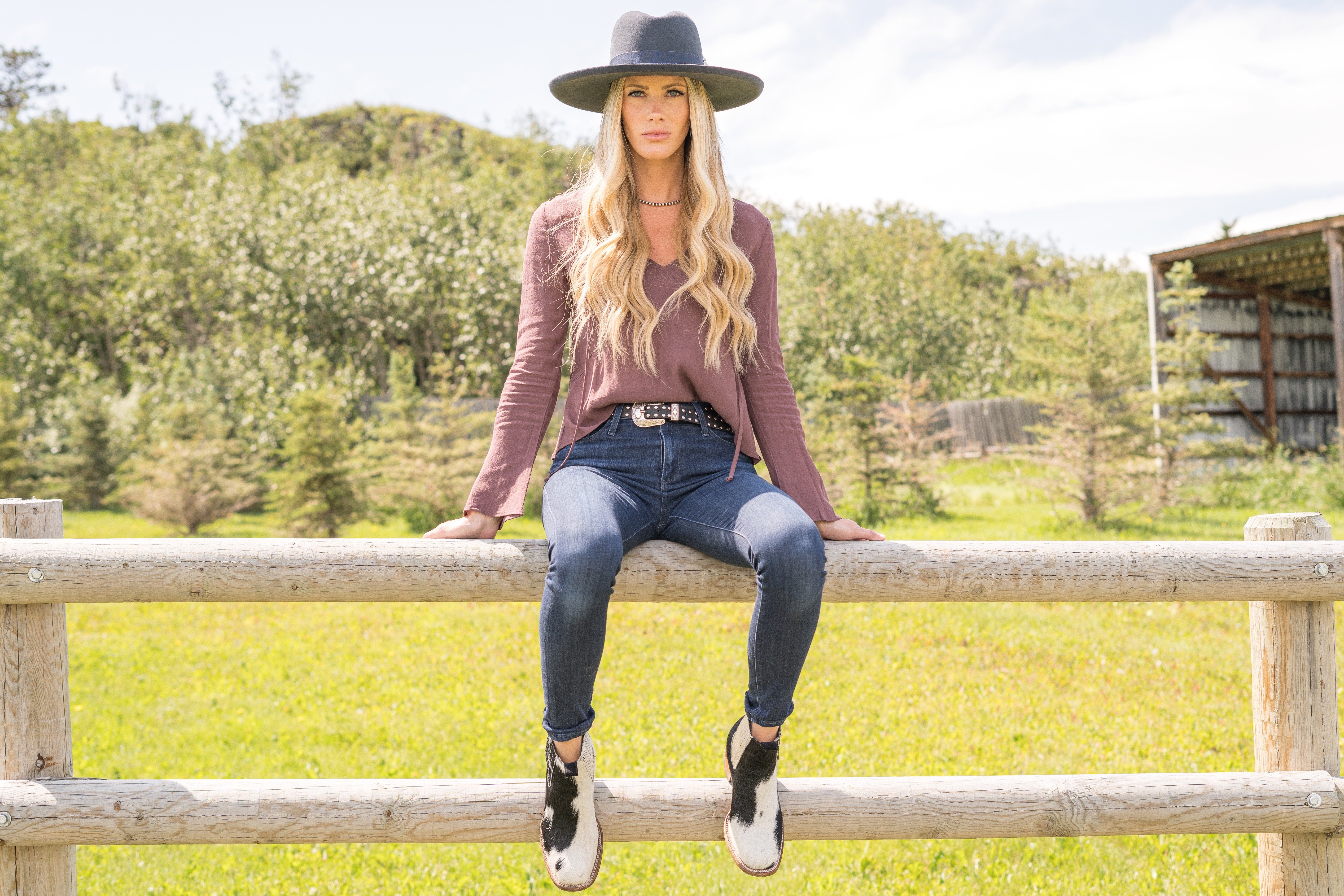 Woman sitting on a wooden fence in a rural setting with green fields and trees wearing cowhide ankle boots